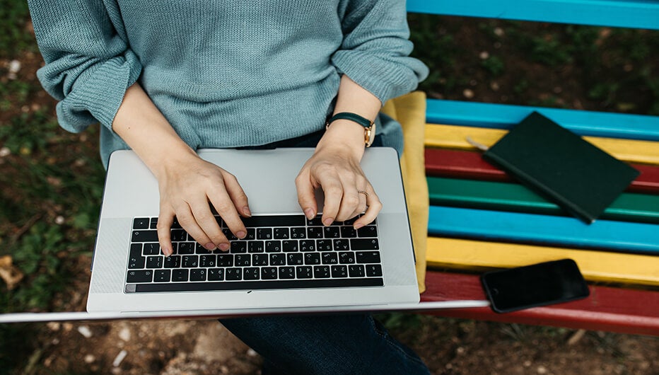 close up of fashionable young Caucasian woman sitting on colourful wooden bench in park. She is keyboarding on laptop. Freelance work concept. Copy space for text. No face shown. 