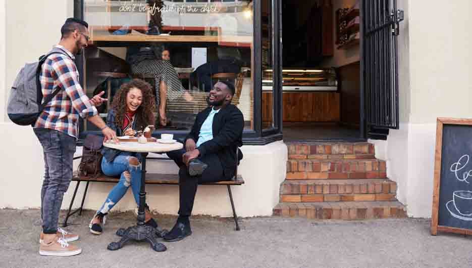Two smiling young people talking and laughing with a friend while sitting at a table at a sidewalk cafe having drinks and cake